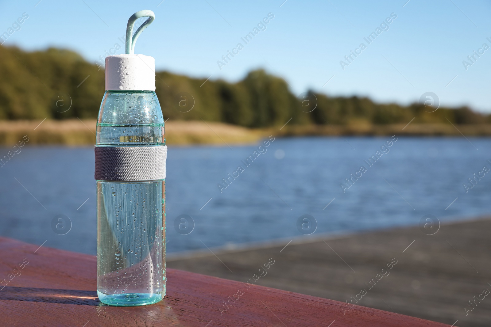 Photo of Glass bottle with water on wooden pier near river outdoors. Space for text