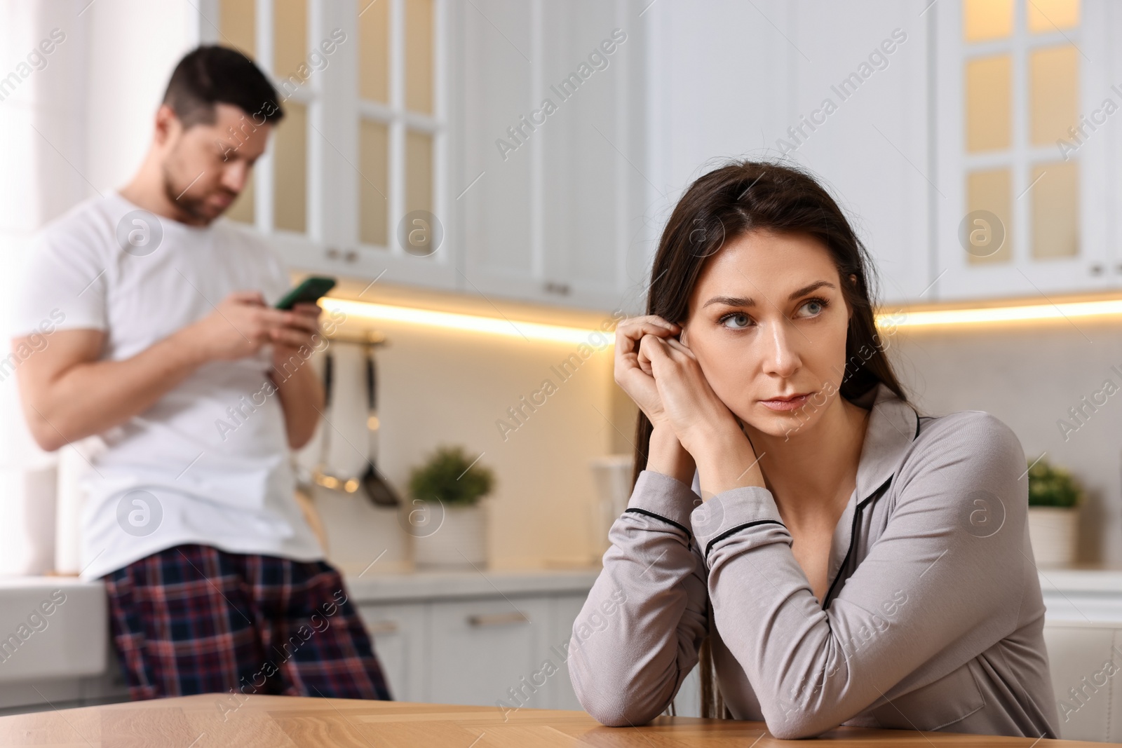 Photo of Offended wife sitting at table while her husband using smartphone in kitchen, selective focus. Relationship problems