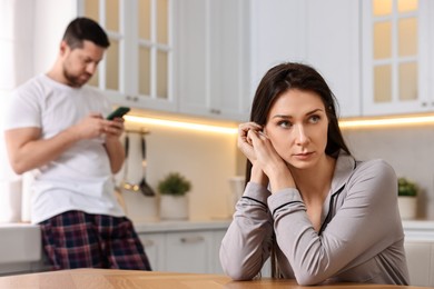 Photo of Offended wife sitting at table while her husband using smartphone in kitchen, selective focus. Relationship problems