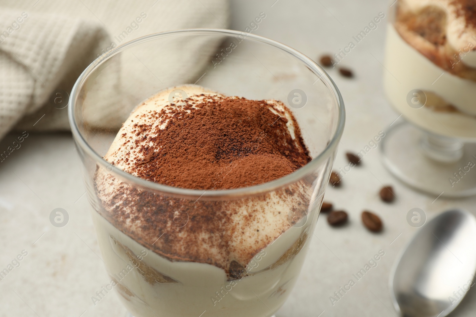 Photo of Delicious tiramisu in glass, spoon and scattered coffee beans on light table, closeup