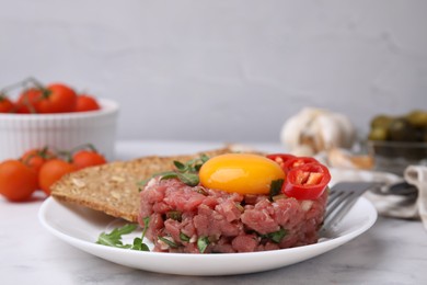 Photo of Tasty beef steak tartare served with yolk, pepper, bread and greens on white marble table