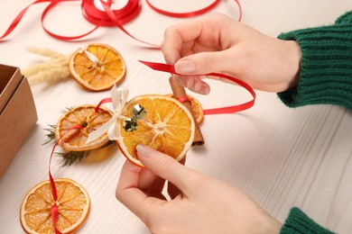 Photo of Woman making garland from dry oranges, cinnamon, ribbon and sleigh bells at white wooden table, closeup