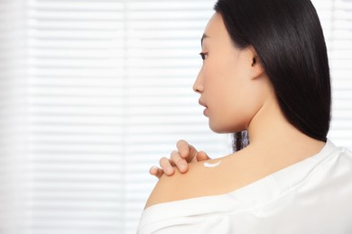 Photo of Beautiful young Asian woman applying body cream on shoulder indoors, space for text