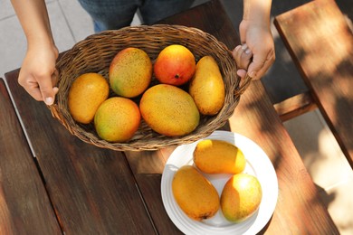 Photo of Woman with wicker basket of tasty mangoes at wooden table, top view