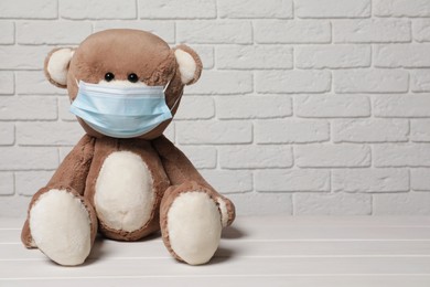 Photo of Cute teddy bear with medical mask on white wooden table near brick wall, space for text