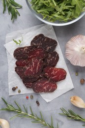 Photo of Slices of delicious beef jerky and spices on grey marble table, flat lay