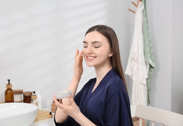 Photo of Beautiful woman in blue robe applying face cream in bathroom
