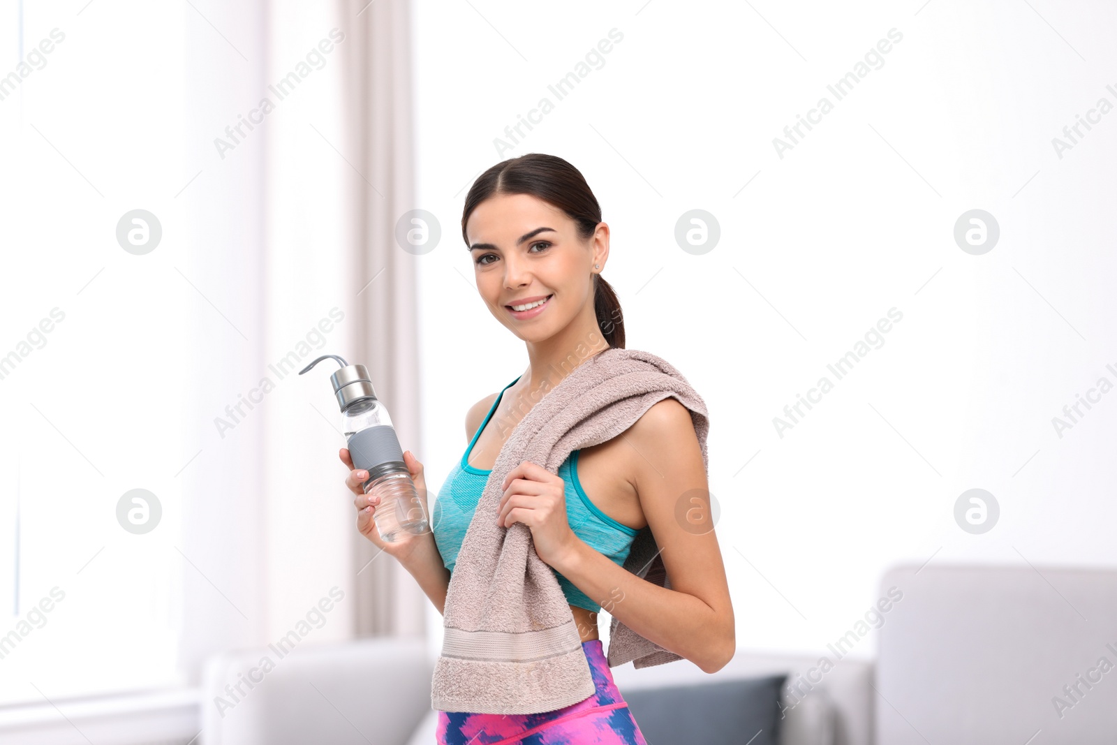 Photo of Beautiful young woman in sportswear with towel and bottle of water indoors