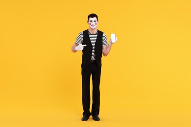 Photo of Funny mime artist pointing at smartphone on orange background