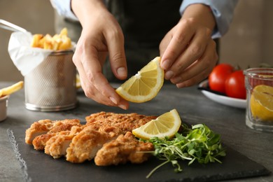Photo of Woman adding lemon onto tasty schnitzels with microgreens at grey table, closeup