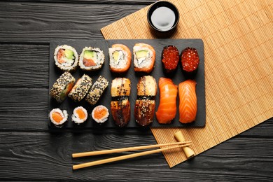 Photo of Delicious sushi rolls, soy sauce and chopsticks on black wooden table, flat lay