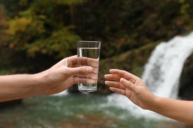 Photo of Man giving woman glass of fresh water near waterfall, closeup
