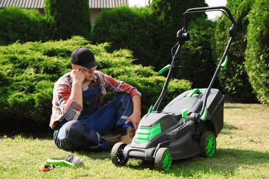 Photo of Tired young man sitting near broken lawn mower in garden