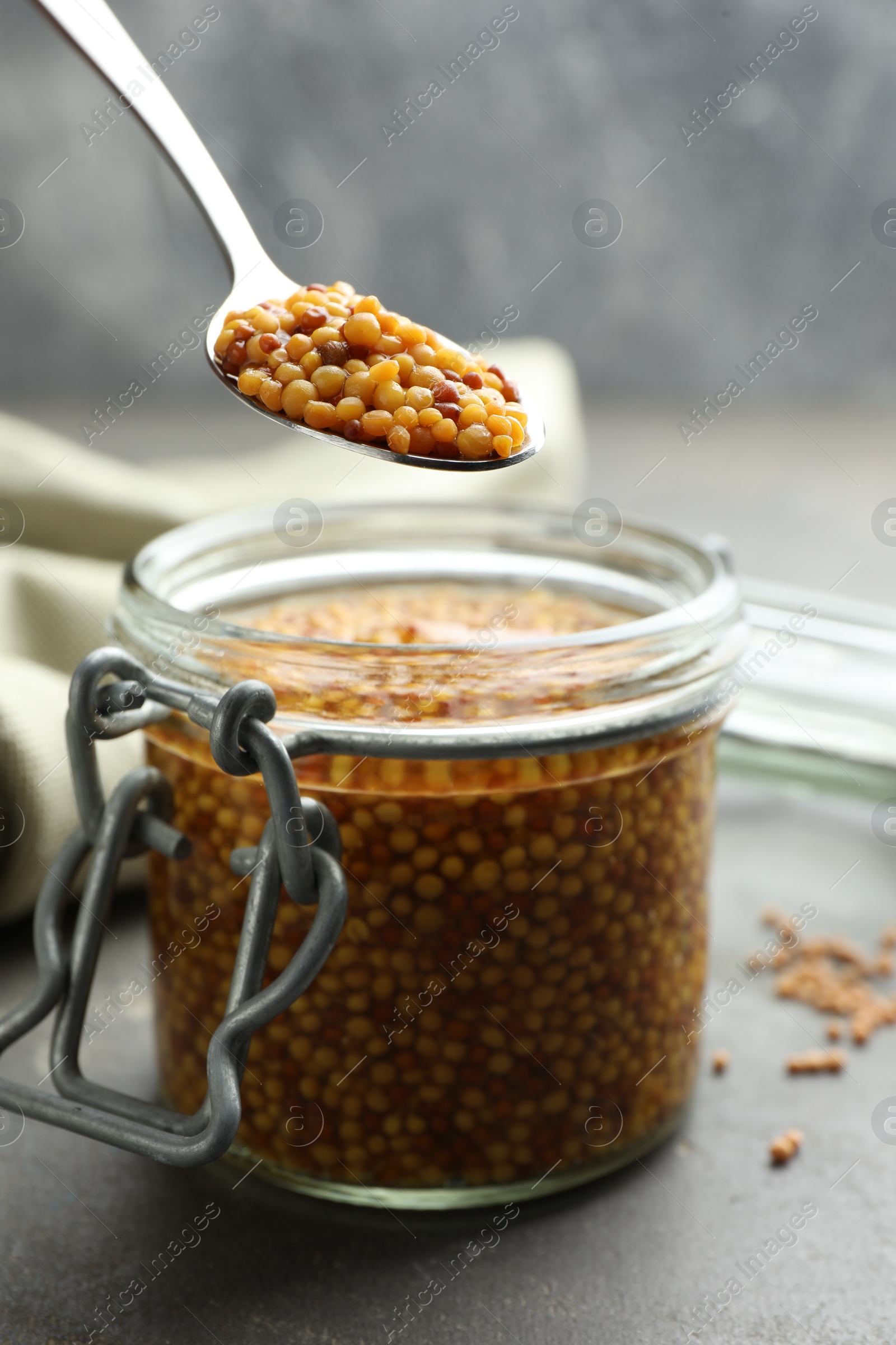 Photo of Taking whole grain mustard with spoon from jar on grey table