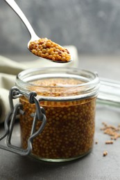 Photo of Taking whole grain mustard with spoon from jar on grey table