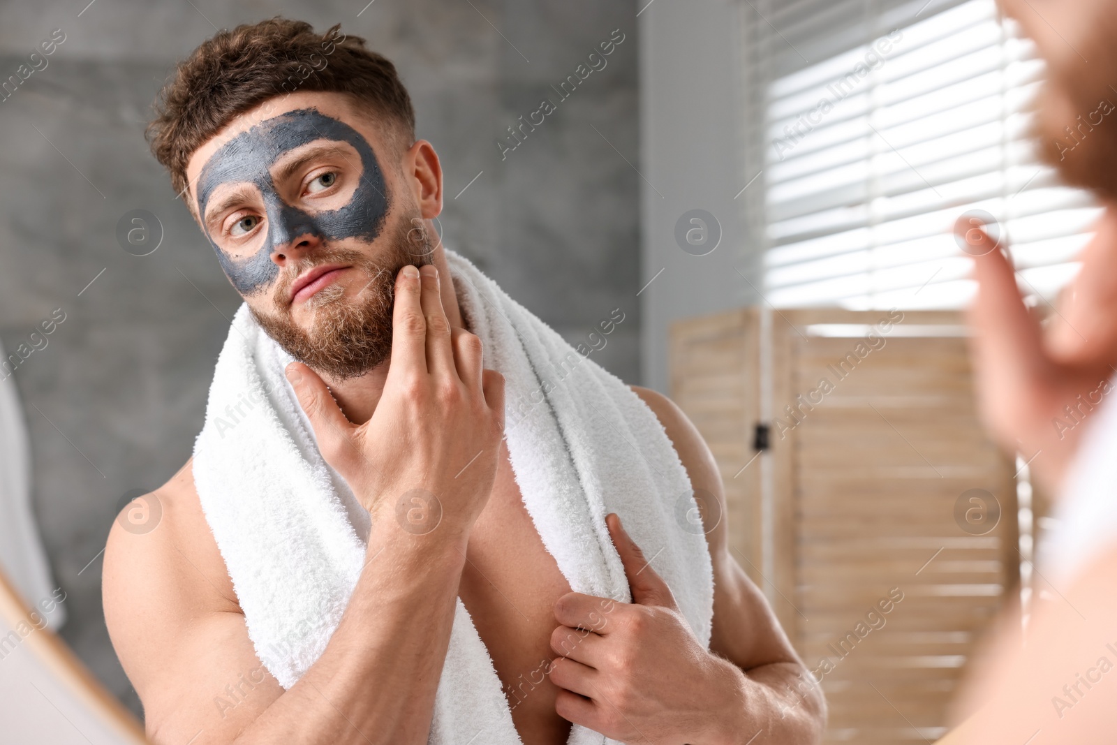 Photo of Handsome man with clay mask on his face near mirror in bathroom