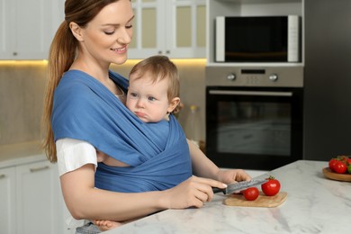 Photo of Mother cutting tomatoes while holding her child in sling (baby carrier) in kitchen