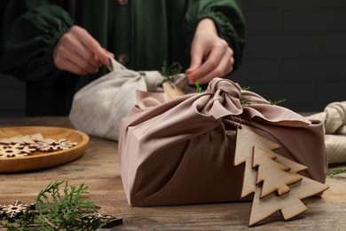 Photo of Wrapped in fabric gift with thuja branches on wooden table. Woman using Furoshiki technique, selective focus