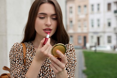 Photo of Beautiful young woman with cosmetic pocket mirror applying lipstick outdoors, space for text