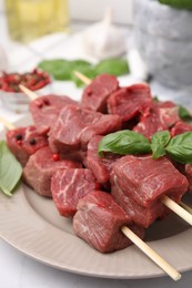 Photo of Wooden skewers with cut fresh beef meat, basil leaves and spices on beige plate, closeup