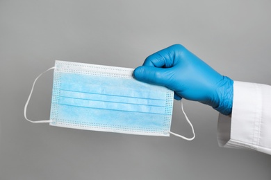 Doctor in medical gloves holding protective mask on light grey background, closeup