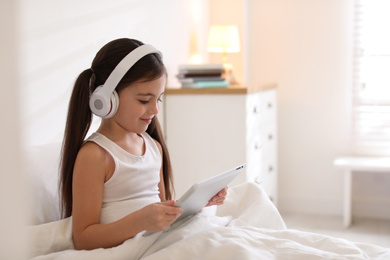 Photo of Cute little girl with headphones and tablet listening to audiobook in bed at home