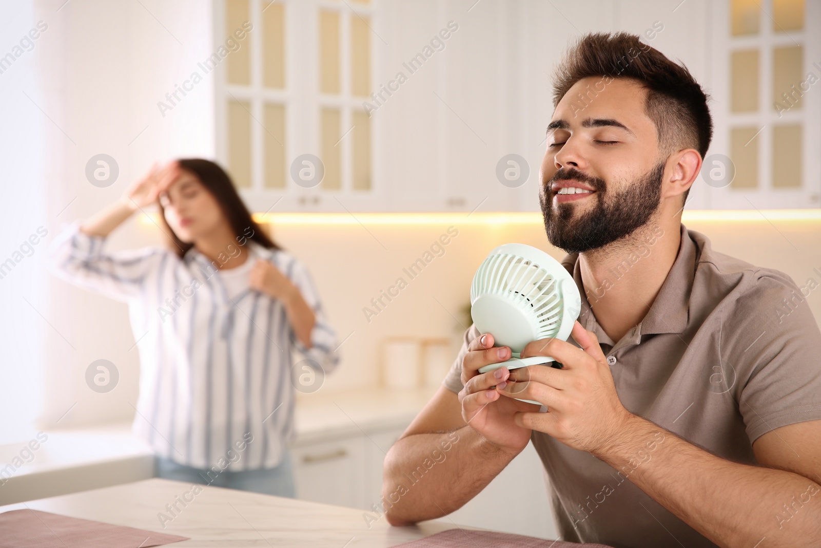 Photo of Man with portable fan enjoying cool air while his girlfriend suffering from heat at home