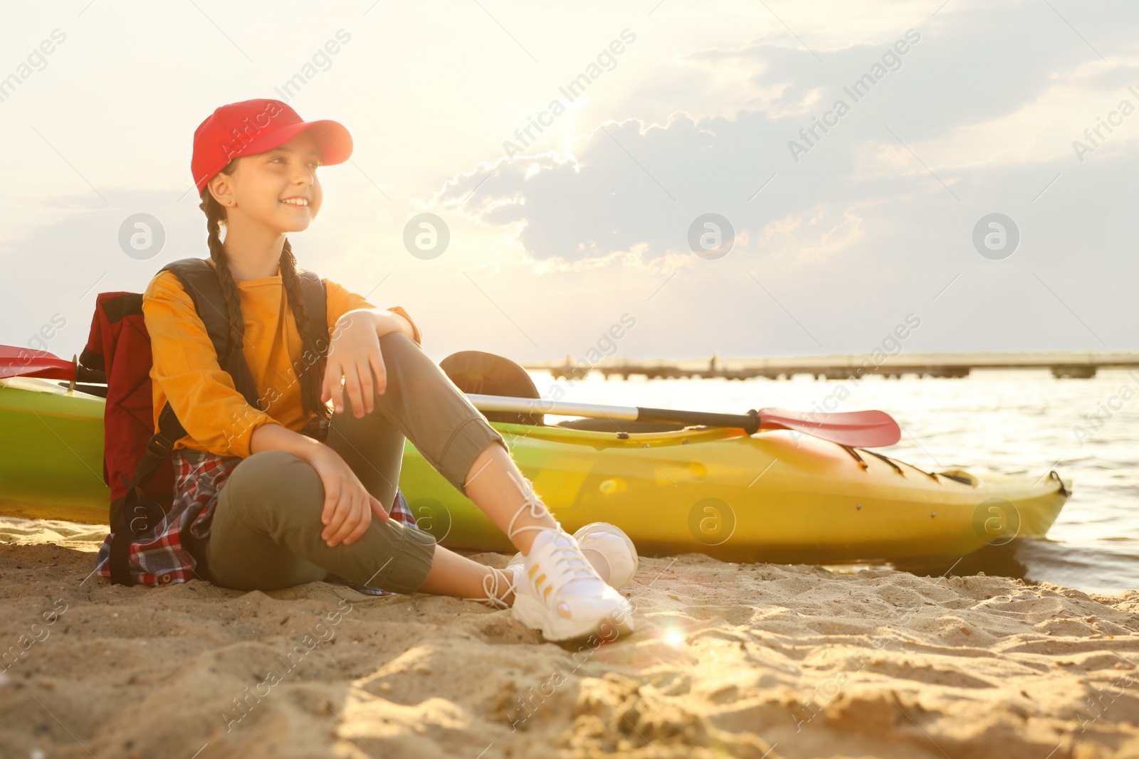 Photo of Happy girl sitting near kayak on river shore. Summer camp activity