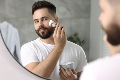 Photo of Handsome young man shaving beard near mirror in bathroom