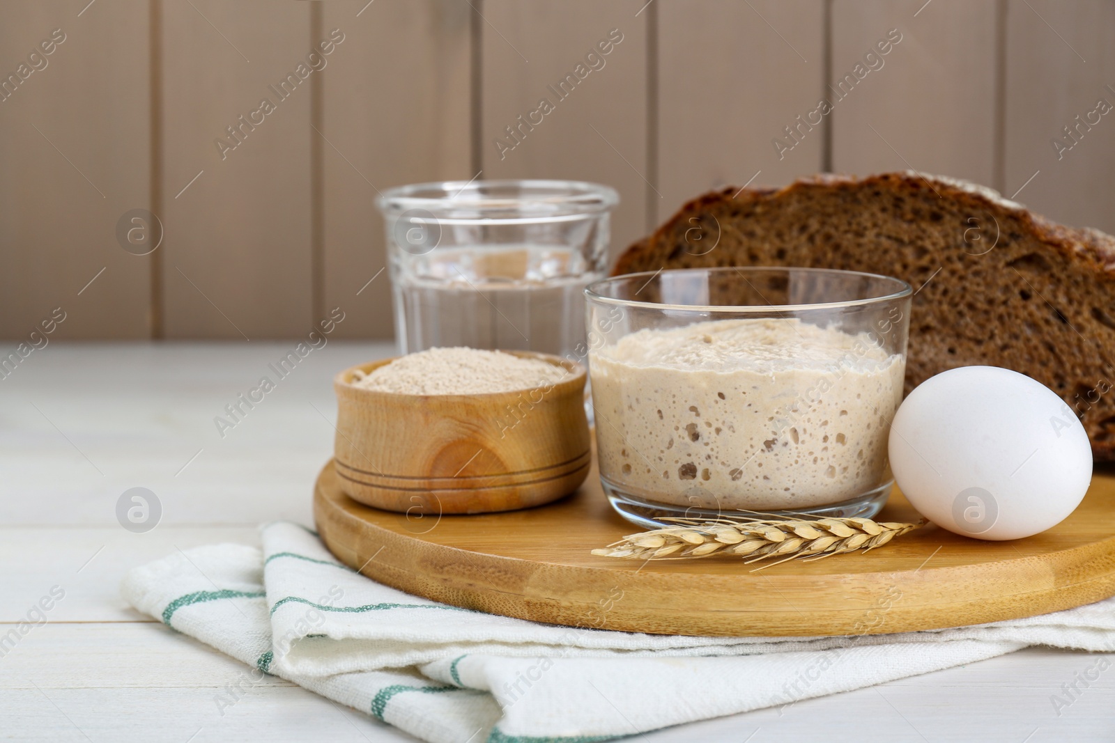 Photo of Glass jar with sourdough and other ingredients for bread on white wooden table