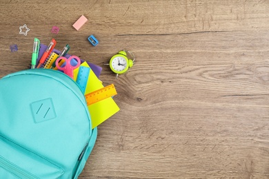 Stylish backpack with different school stationary and alarm clock on wooden table, flat lay. Space for text