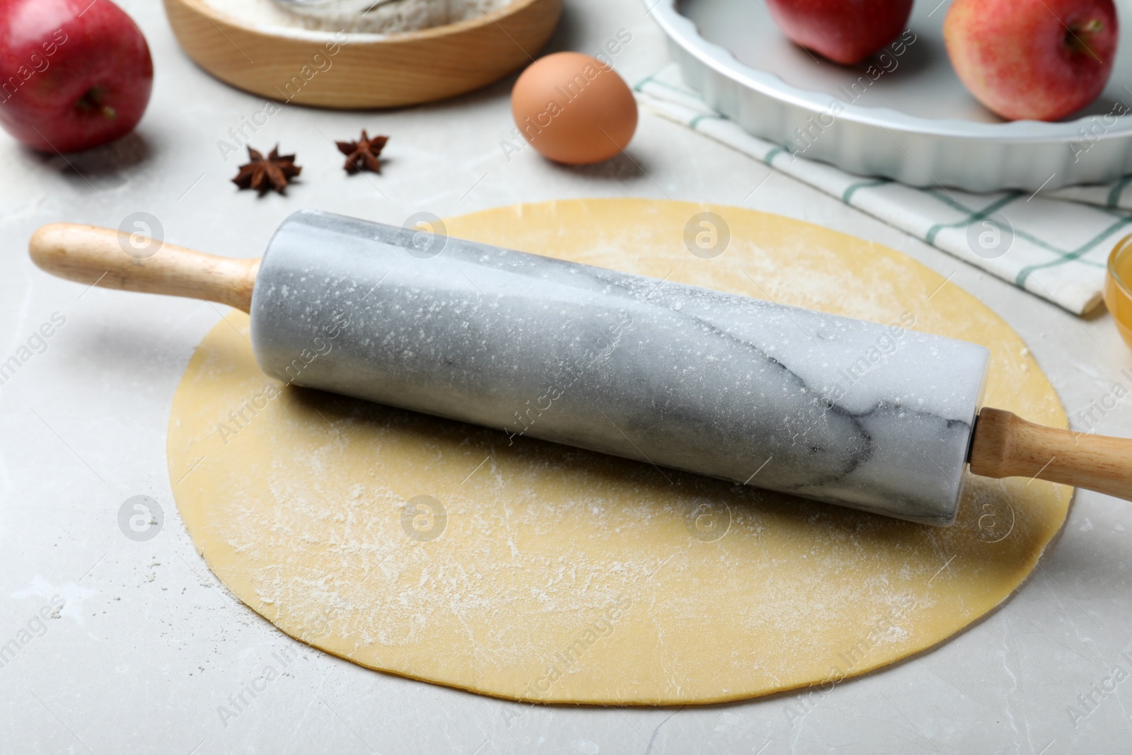Photo of Rolling pin, raw dough and ingredients on light grey table. Baking apple pie