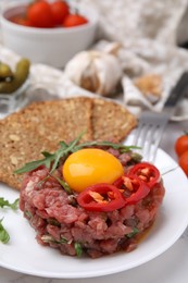 Photo of Tasty beef steak tartare served with yolk, pepper, bread and greens on white table, closeup