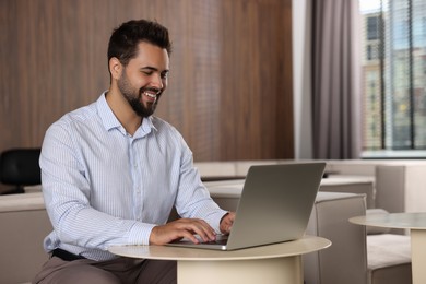 Happy young man working on laptop at table in office. Space for text