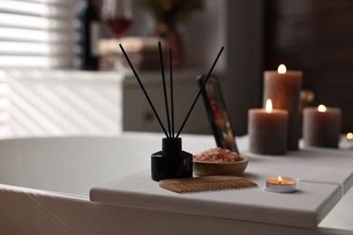 Photo of White wooden tray with burning candles, aroma diffuser and sea salt on bathtub in bathroom, space for text