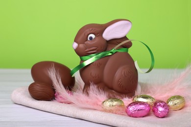 Chocolate Easter bunny, eggs and feathers on white wooden table against light green background, closeup