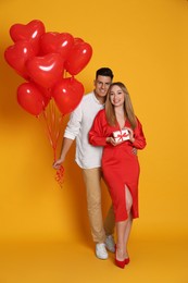 Photo of Lovely couple with heart shaped balloons and gift on yellow background. Valentine's day celebration