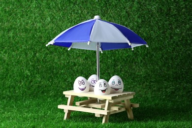 Photo of Eggs with drawn happy faces, small picnic table and umbrella on green grass