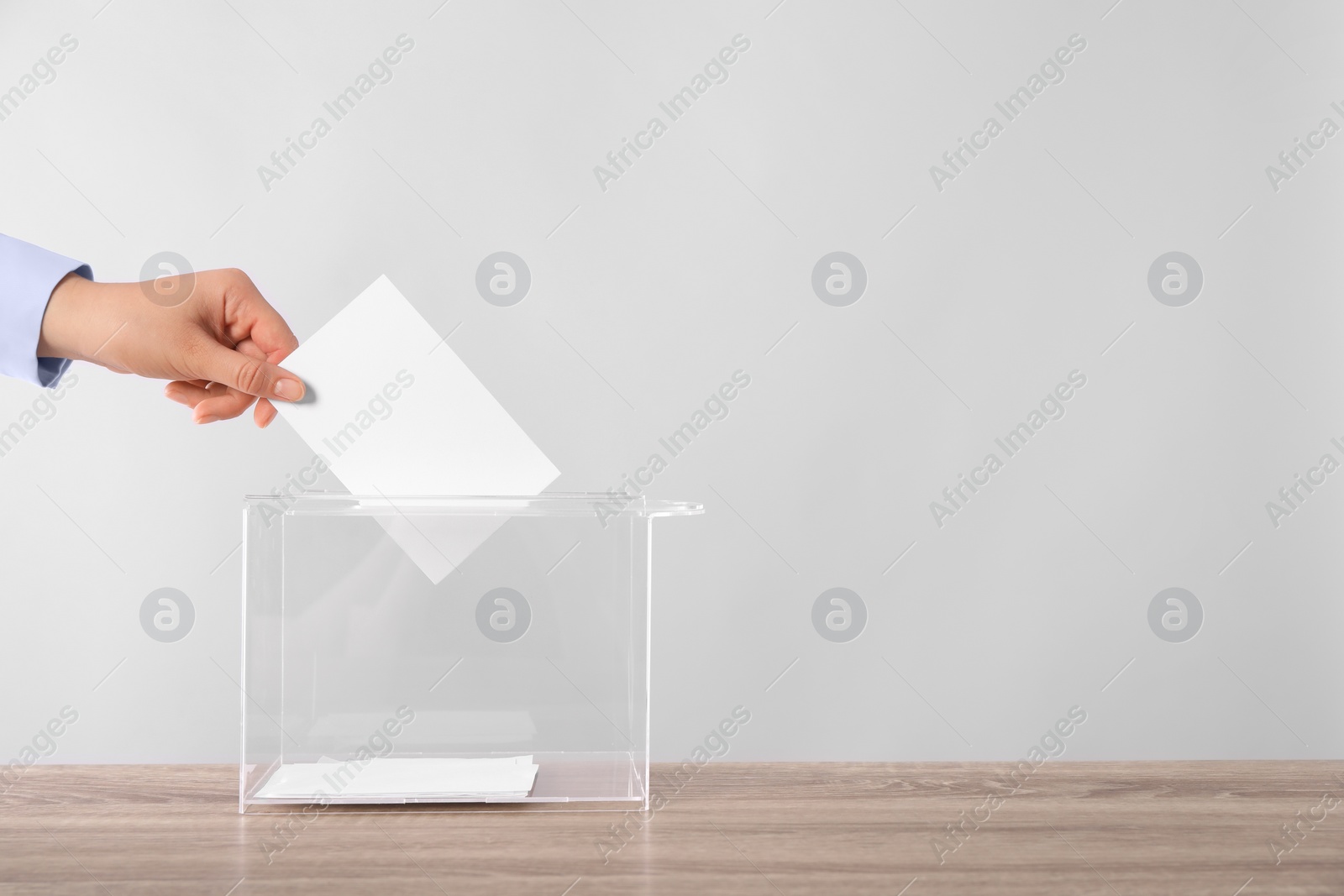 Photo of Woman putting her vote into ballot box on wooden table against light grey background, closeup. Space for text