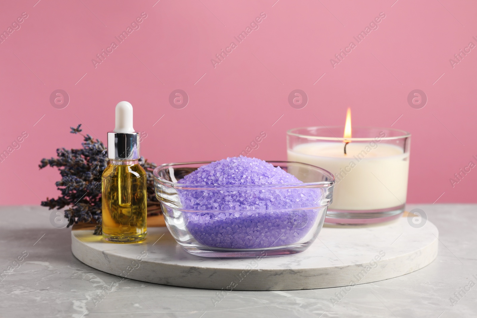 Photo of Violet sea salt in bowl, lavender flowers, burning candle and cosmetic product on grey marble table