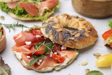 Photo of Tasty bagel with cured ham, cheese, tomatoes and arugula on white tiled table, closeup