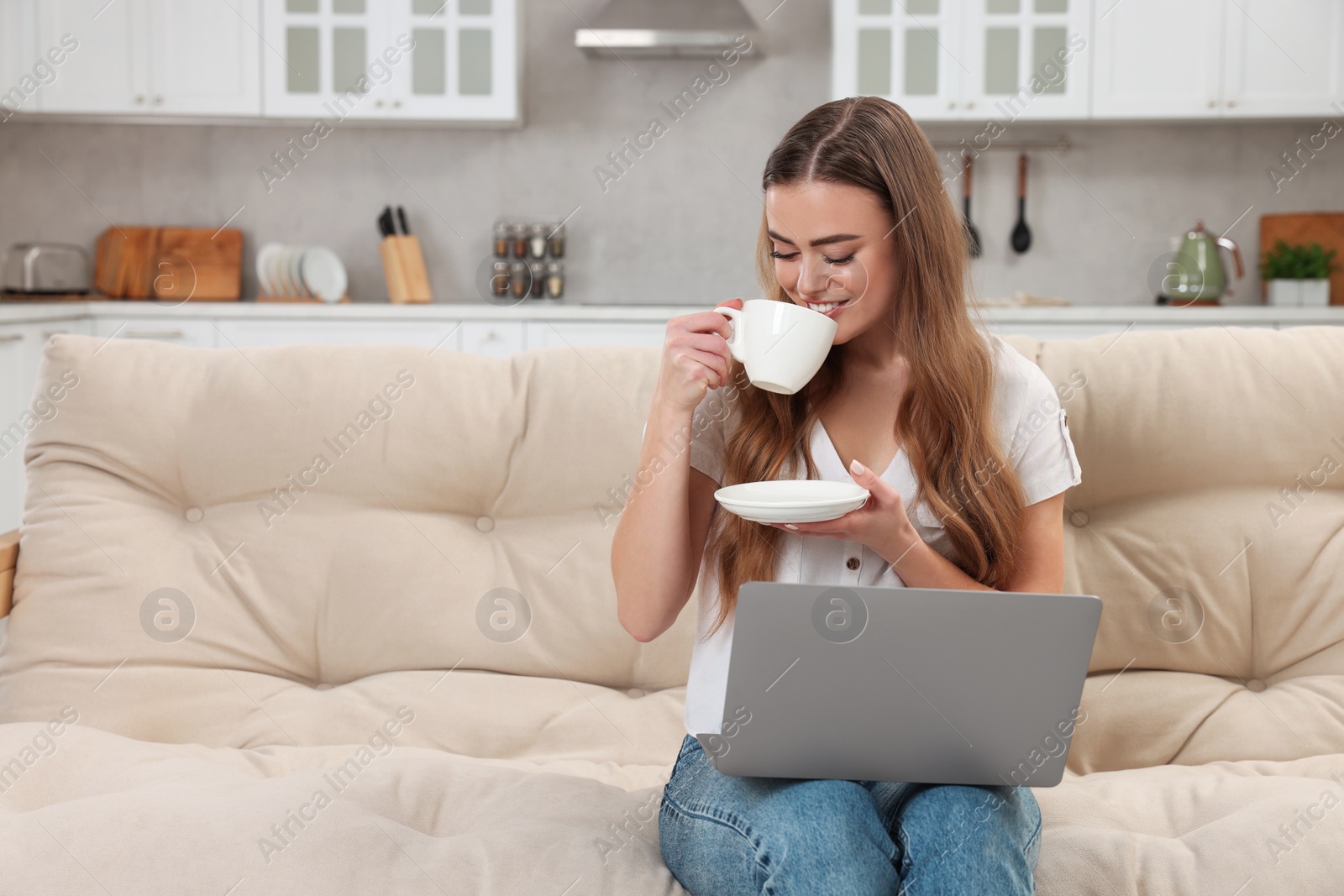 Photo of Happy woman drinking coffee near laptop on couch in room