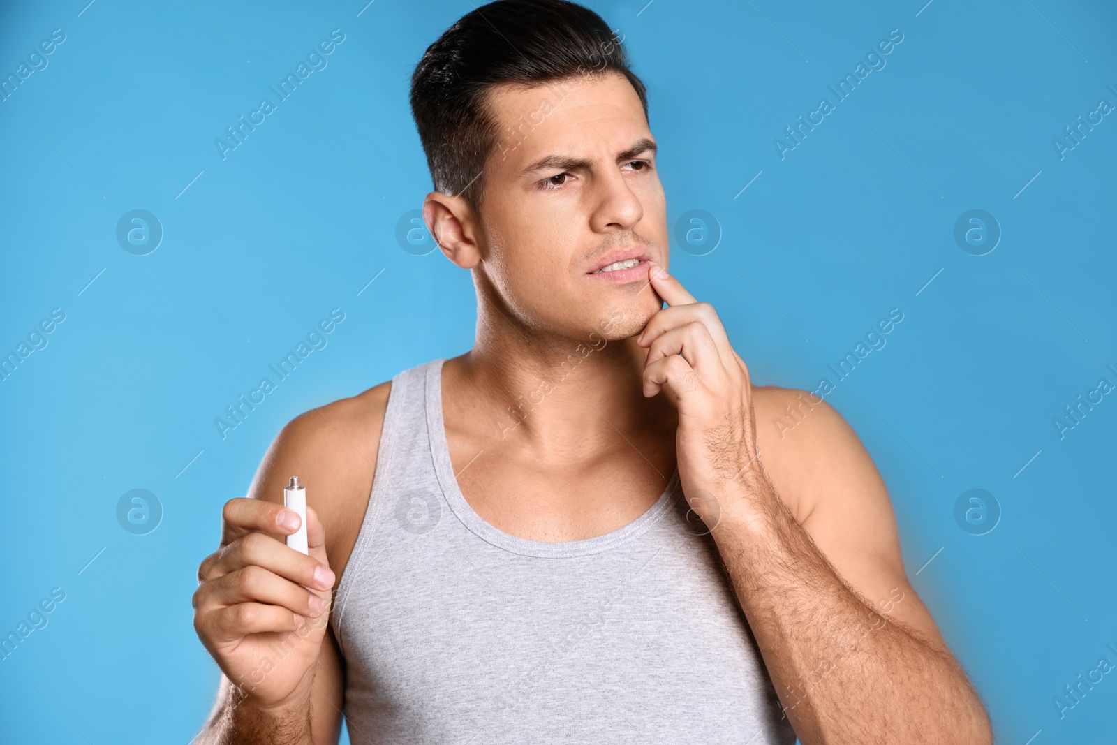 Photo of Emotional man with herpes applying cream on lips against light blue background