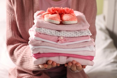Photo of Woman holding stack of baby girl's clothes with shoes indoors, closeup