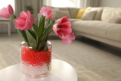 Photo of Different color fillers and tulips in glass vase on white table at home, space for text. Water beads