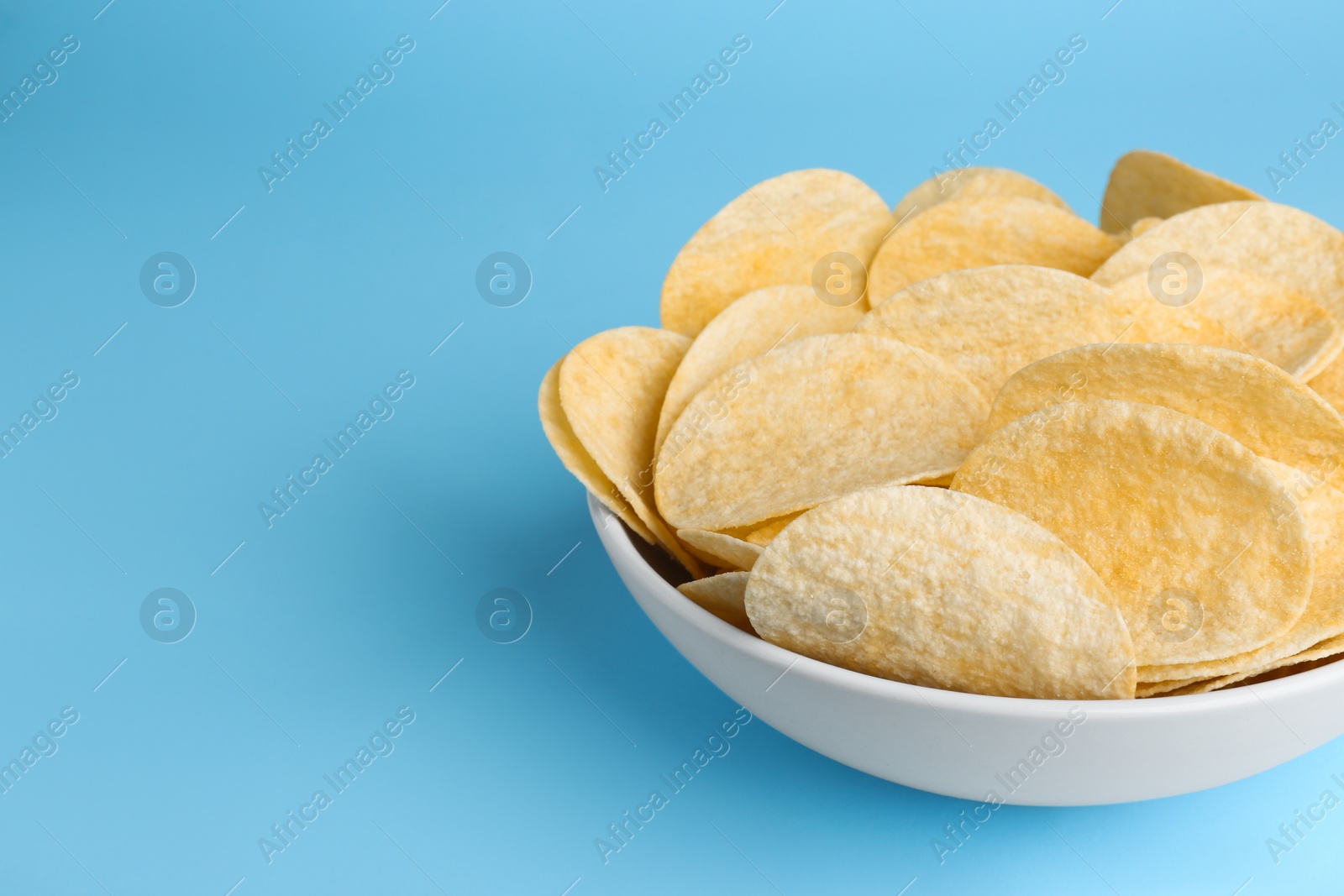 Photo of Bowl of tasty potato chips on light blue background, closeup. Space for text