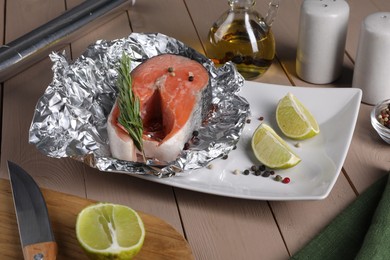 Photo of Aluminum foil with raw salmon, lime slices, rosemary and spices on wooden table
