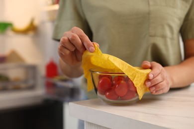 Photo of Woman covering bowl of fresh tomatoes with beeswax food wrap at table in kitchen, closeup. Space for text