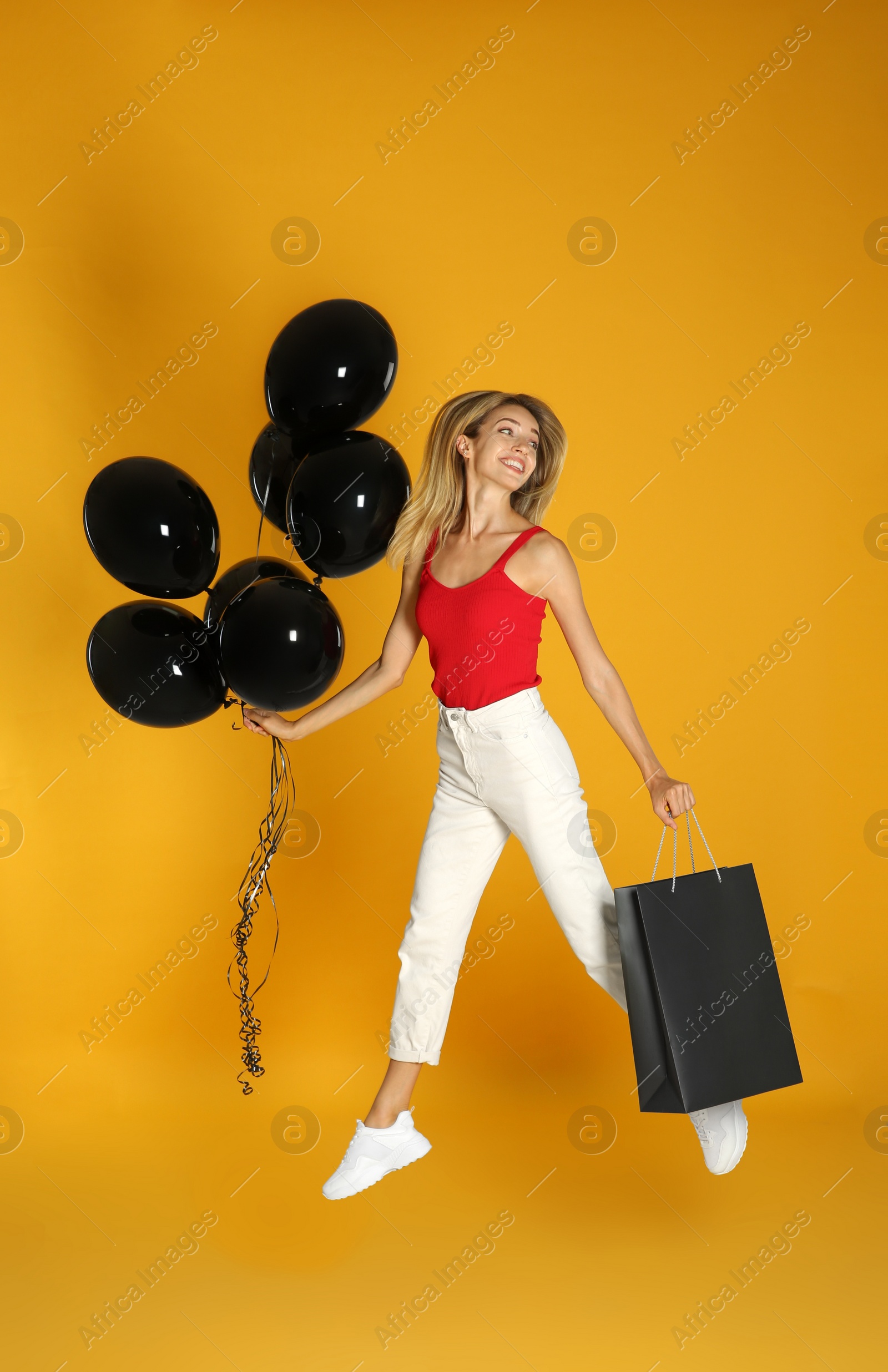Photo of Happy young woman with balloons and shopping bag jumping on yellow background. Black Friday Sale