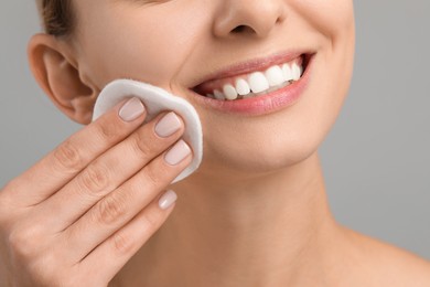 Photo of Smiling woman removing makeup with cotton pad on grey background, closeup